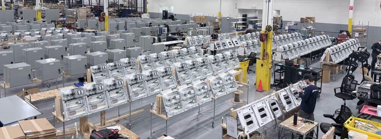 Panel shop floor with rows of open control panels on workbenches and gray enclosures, with technicians assembling them