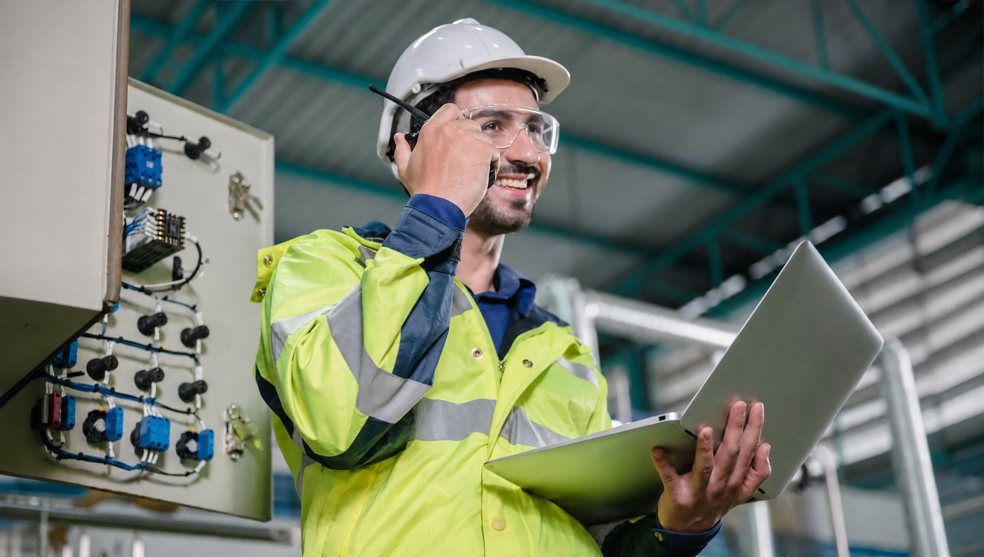 A field engineer in a hard hat and high-visibility jacket holding a laptop and radio beside an open electrical control panel